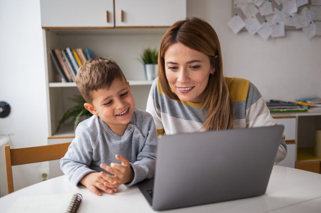 A woman and a young boy sit at a table, smiling and looking at a laptop screen together in a cozy, well-lit room with shelves and papers on the wall in the background.