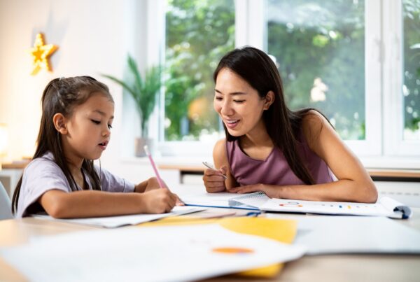A woman and a young girl sit at a table by a window, working together on schoolwork or a drawing. The woman is smiling and holding a pen, while the girl writes with a pencil. Papers and notebooks are spread out on the table.