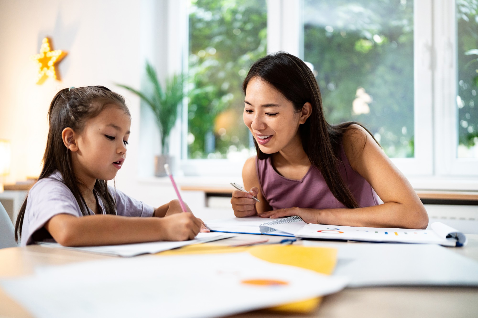 A woman and a young girl sit at a table by a window, working together on schoolwork or a drawing. The woman is smiling and holding a pen, while the girl writes with a pencil. Papers and notebooks are spread out on the table.