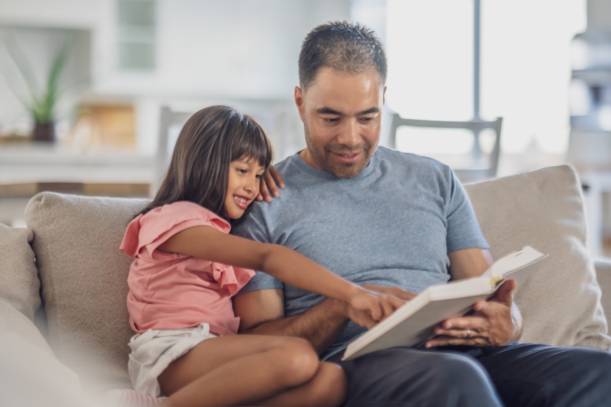 A young girl and an adult man sit together on a couch, smiling and reading a book. The girl is pointing at something on the page while the man holds the book and looks on, both appearing happy and engaged.