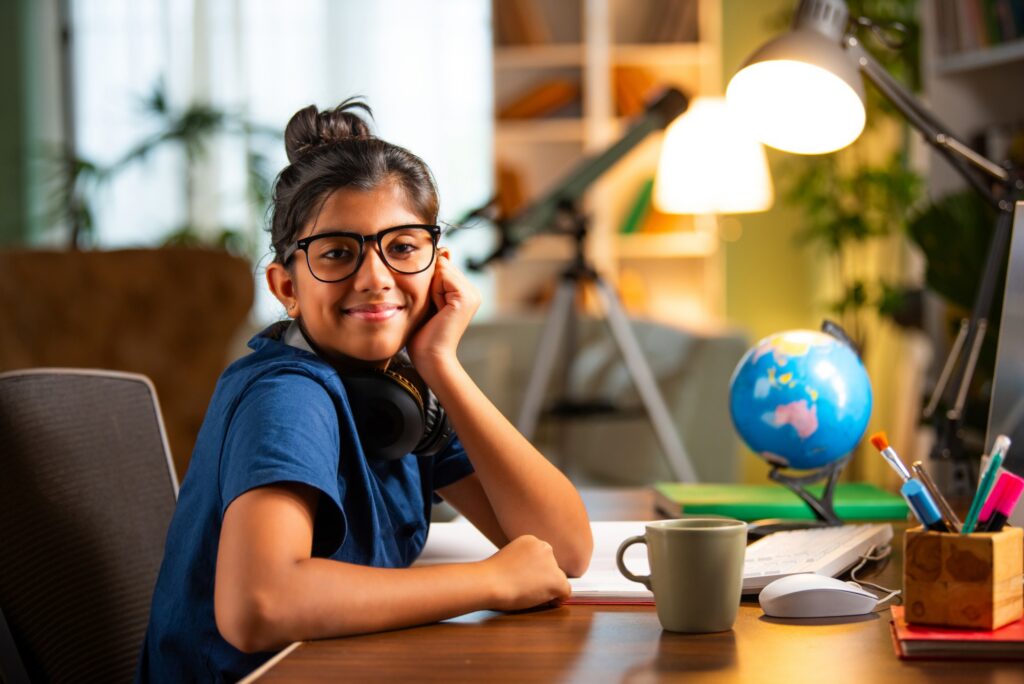 A smiling girl with glasses and headphones sits at a desk with a mug, globe, and stationery. Background shows bookshelves, plants, a lamp, and a telescope, creating a cozy study space.