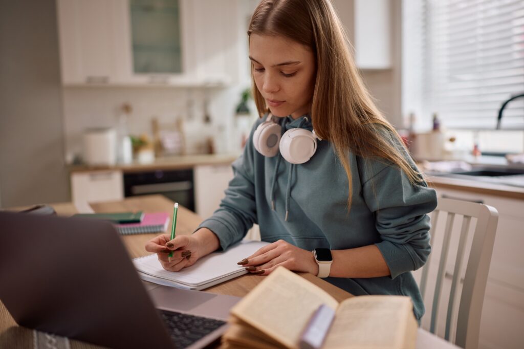 A young woman with headphones around her neck writes in a notebook while studying at a kitchen table with an open laptop and book in front of her. She appears focused and is surrounded by study materials.