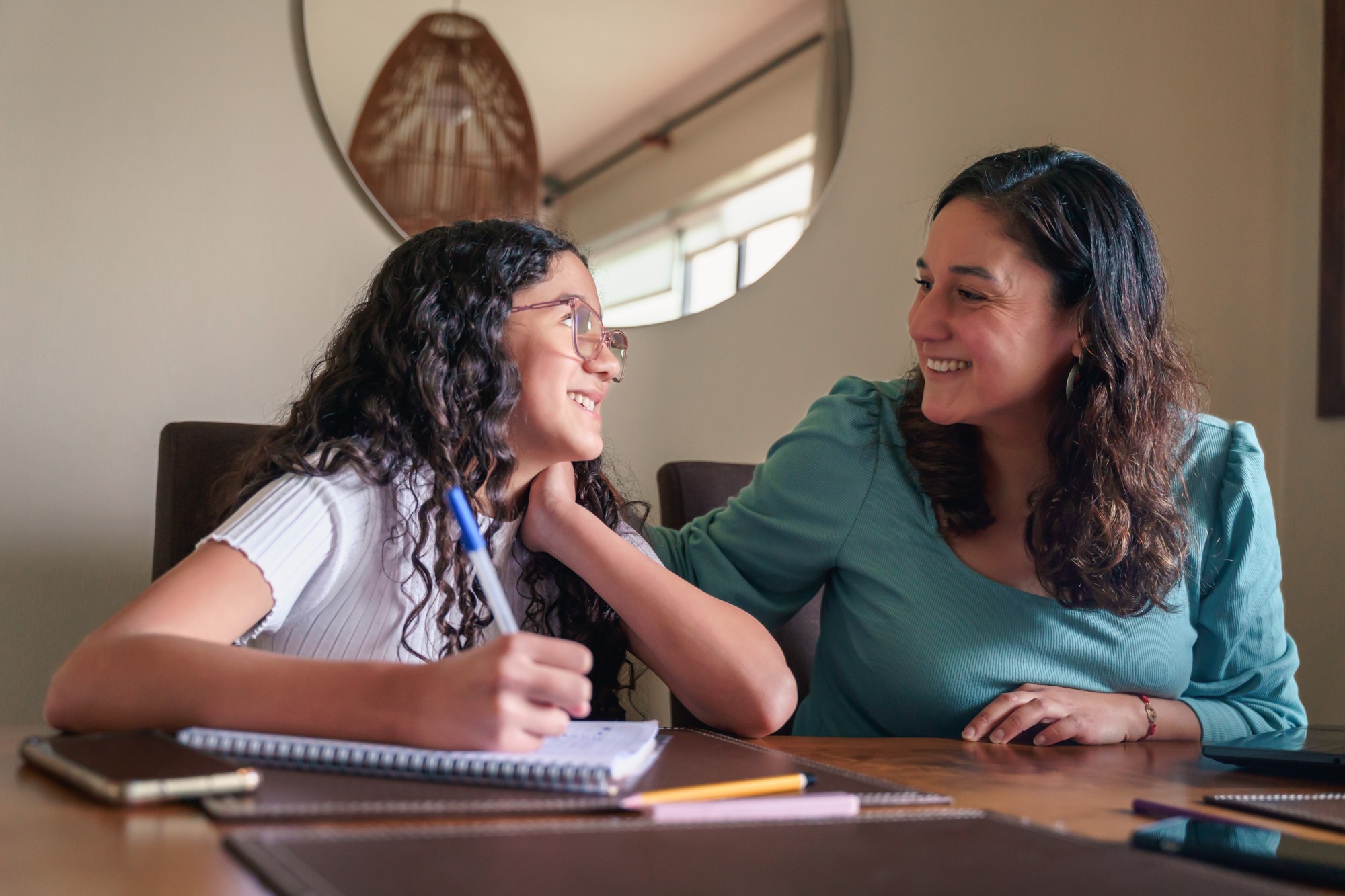 A woman and a girl with curly hair sit at a table, smiling at each other. The girl holds a pen over a notebook, while the woman rests her hand on the girls shoulder. They appear to be working or studying together.