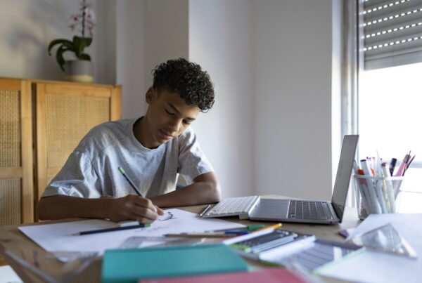 A teenage boy sits at a desk drawing on paper, with colored pencils and art supplies scattered around. An open laptop is nearby, and sunlight filters through a window beside him.