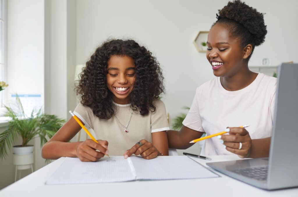A young girl and an adult woman sit at a table, smiling while working together on schoolwork. They both hold pencils and have notebooks open, with a laptop and houseplants in the background.