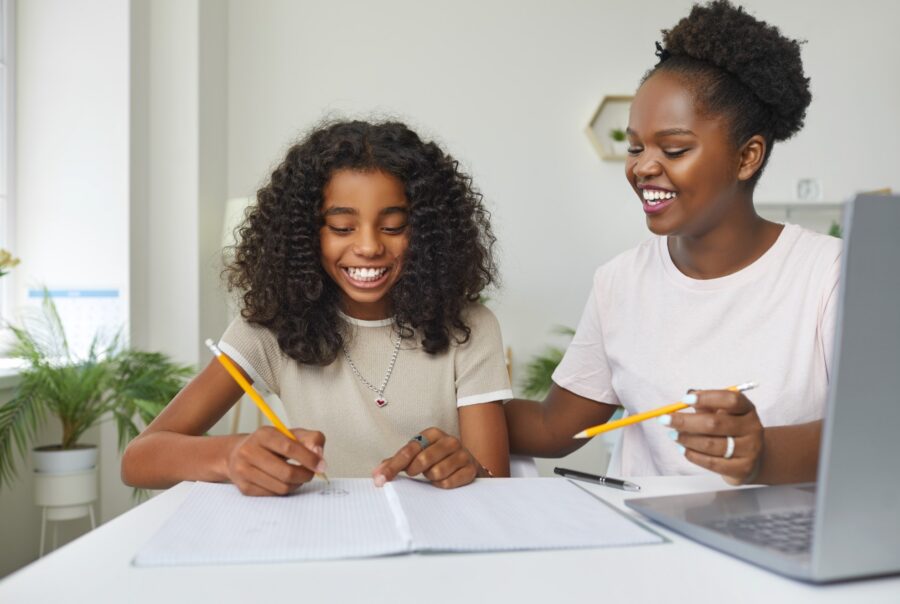 A young girl and an adult woman sit at a table, smiling while working together on schoolwork. They both hold pencils and have notebooks open, with a laptop and houseplants in the background.
