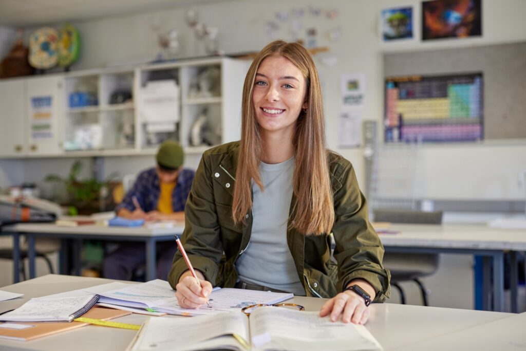 A young woman sits at a desk in a classroom, smiling at the camera while writing in a notebook. Open books, papers, and a pencil are in front of her. Another student is blurred in the background.