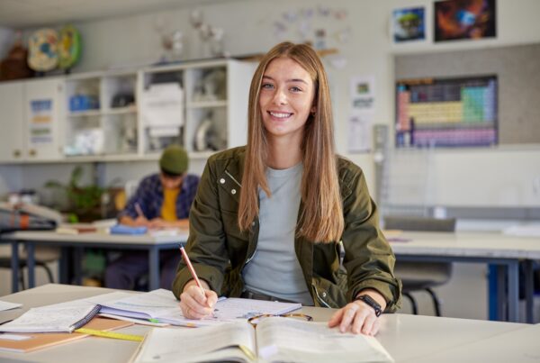 A young woman sits at a desk in a classroom, smiling at the camera while writing in a notebook. Open books, papers, and a pencil are in front of her. Another student is blurred in the background.