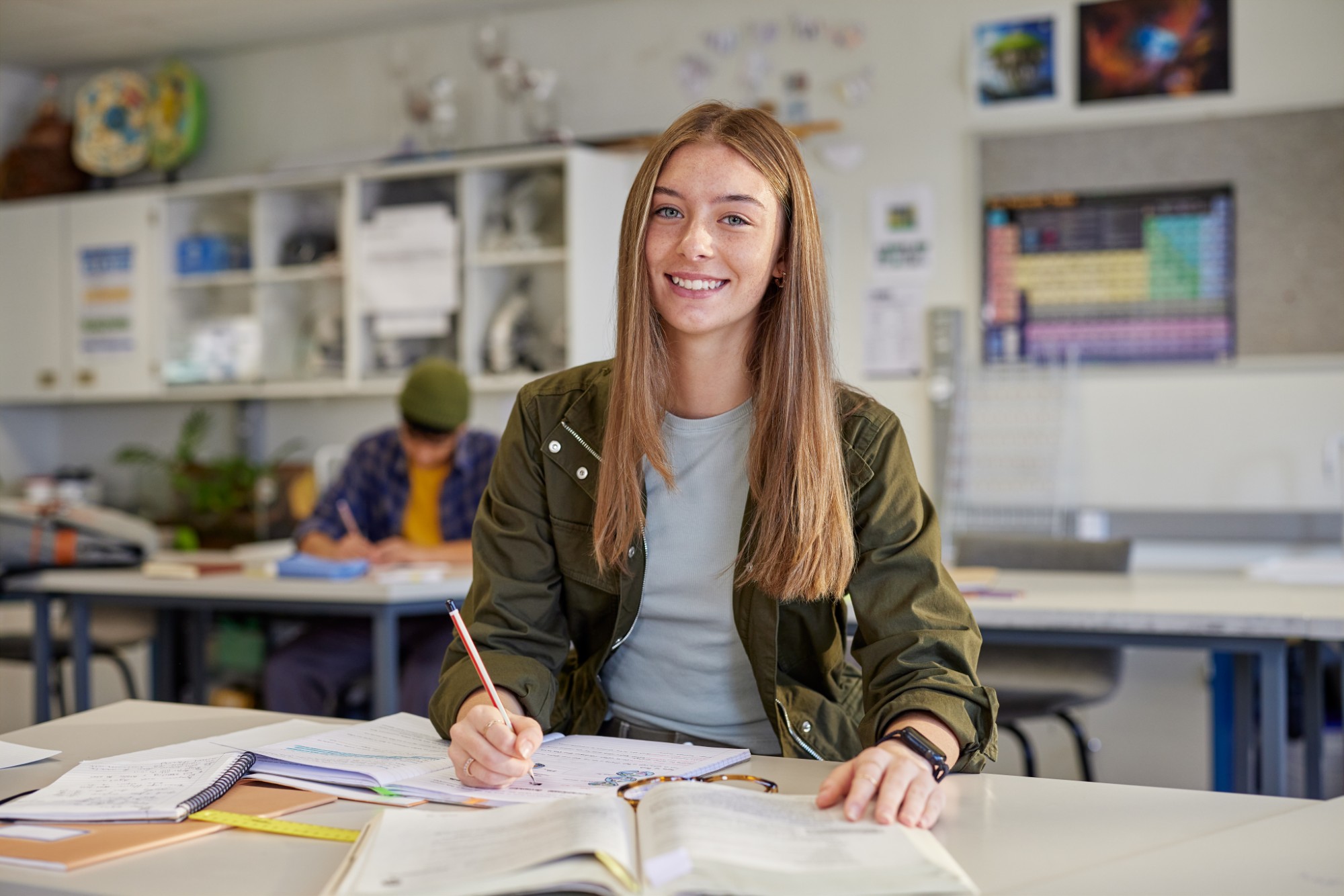 A young woman sits at a desk in a classroom, smiling at the camera while writing in a notebook. Open books, papers, and a pencil are in front of her. Another student is blurred in the background.