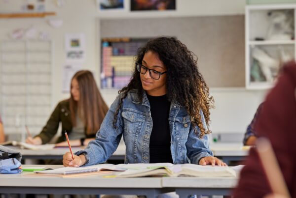 A student with curly hair and glasses, wearing a denim jacket, sits at a desk in a classroom writing in a notebook with open books in front of her. Other students are also seated and working in the background.