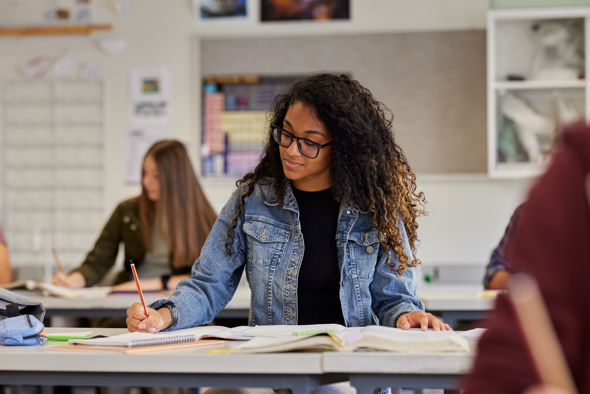 A student with curly hair and glasses, wearing a denim jacket, sits at a desk in a classroom writing in a notebook with open books in front of her. Other students are also seated and working in the background.