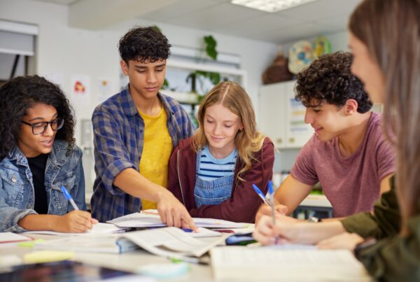 A group of five students sits around a table, studying together with open books and notebooks. One student points at a book while others listen and engage, collaborating in a bright classroom setting.