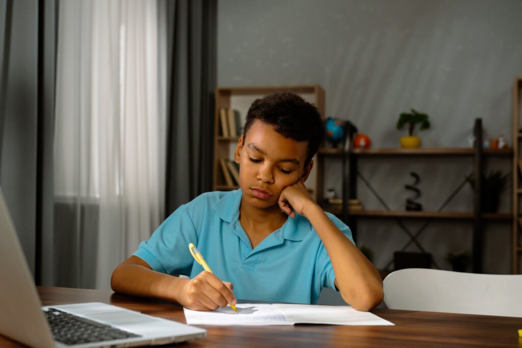 A young boy in a blue shirt sits at a table, resting his head on one hand while writing in a notebook, appearing focused. A laptop and books are on the table, with shelves and a window in the background.