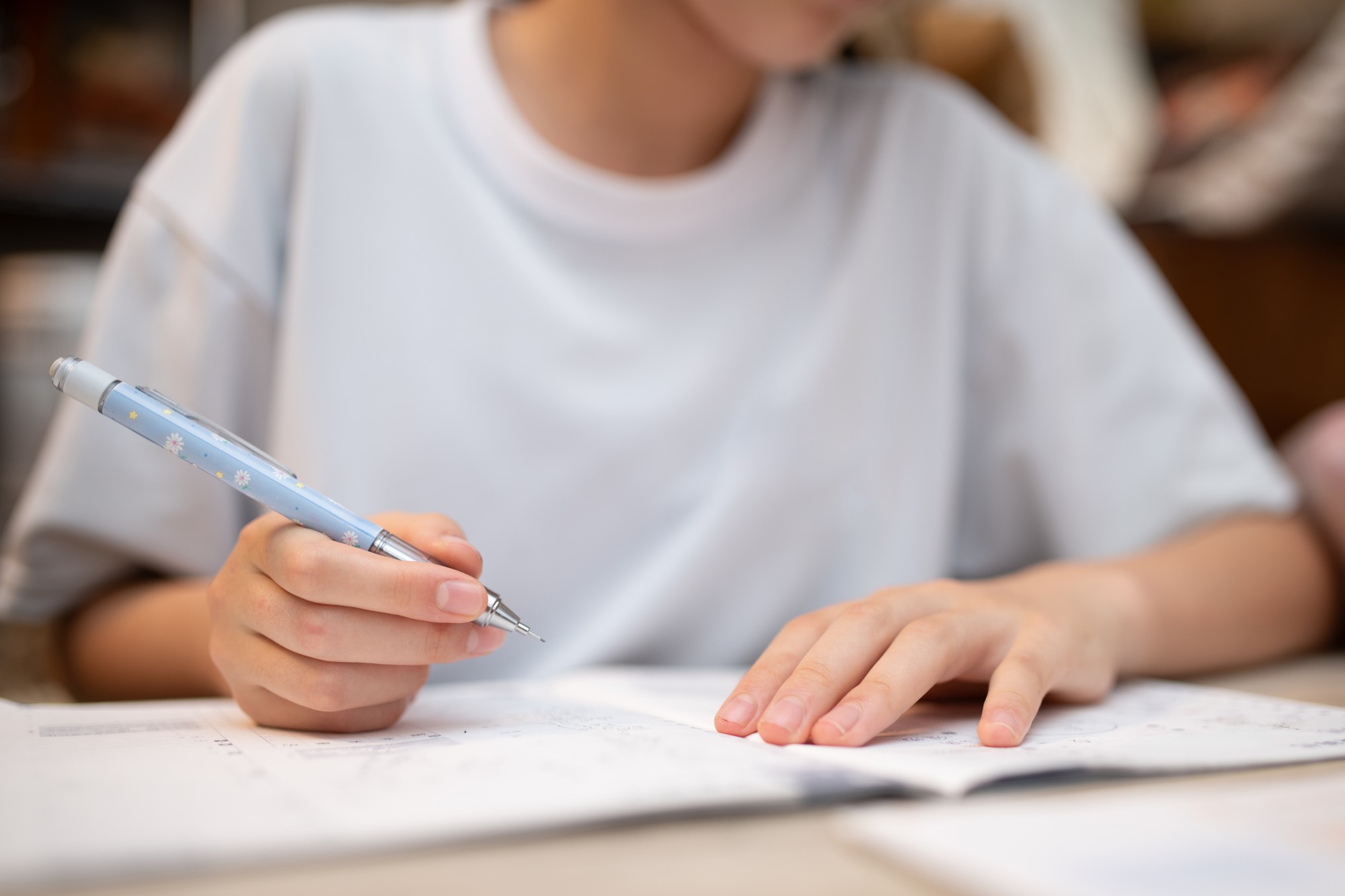 A person wearing a white shirt is writing with a blue pen while studying or doing homework, with an open notebook or workbook in front of them. The focus is on their hands and the pen.