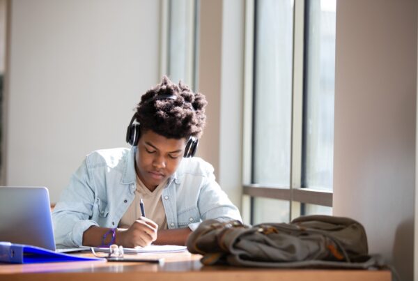 A student wearing headphones sits at a desk by a window, writing in a notebook. A laptop, a blue folder, and a backpack are on the desk beside him. Sunlight streams in through the large windows.