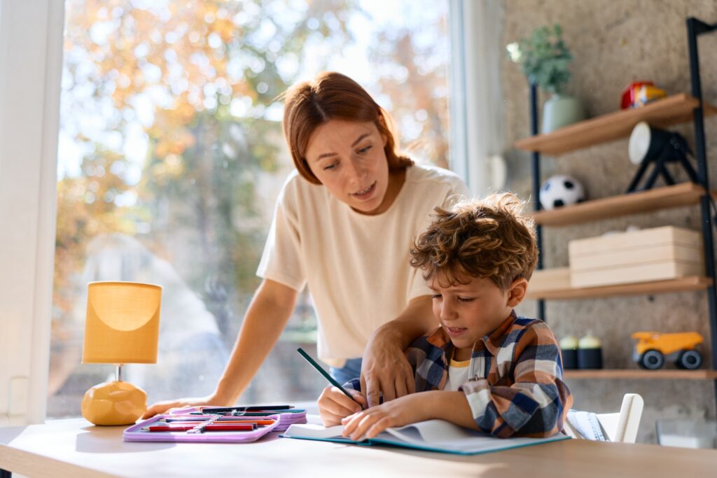 A woman helps a young boy with his homework at a desk by a window. Art supplies and a lamp are on the desk, and shelves with toys are seen in the background. Sunlight streams into the room.
