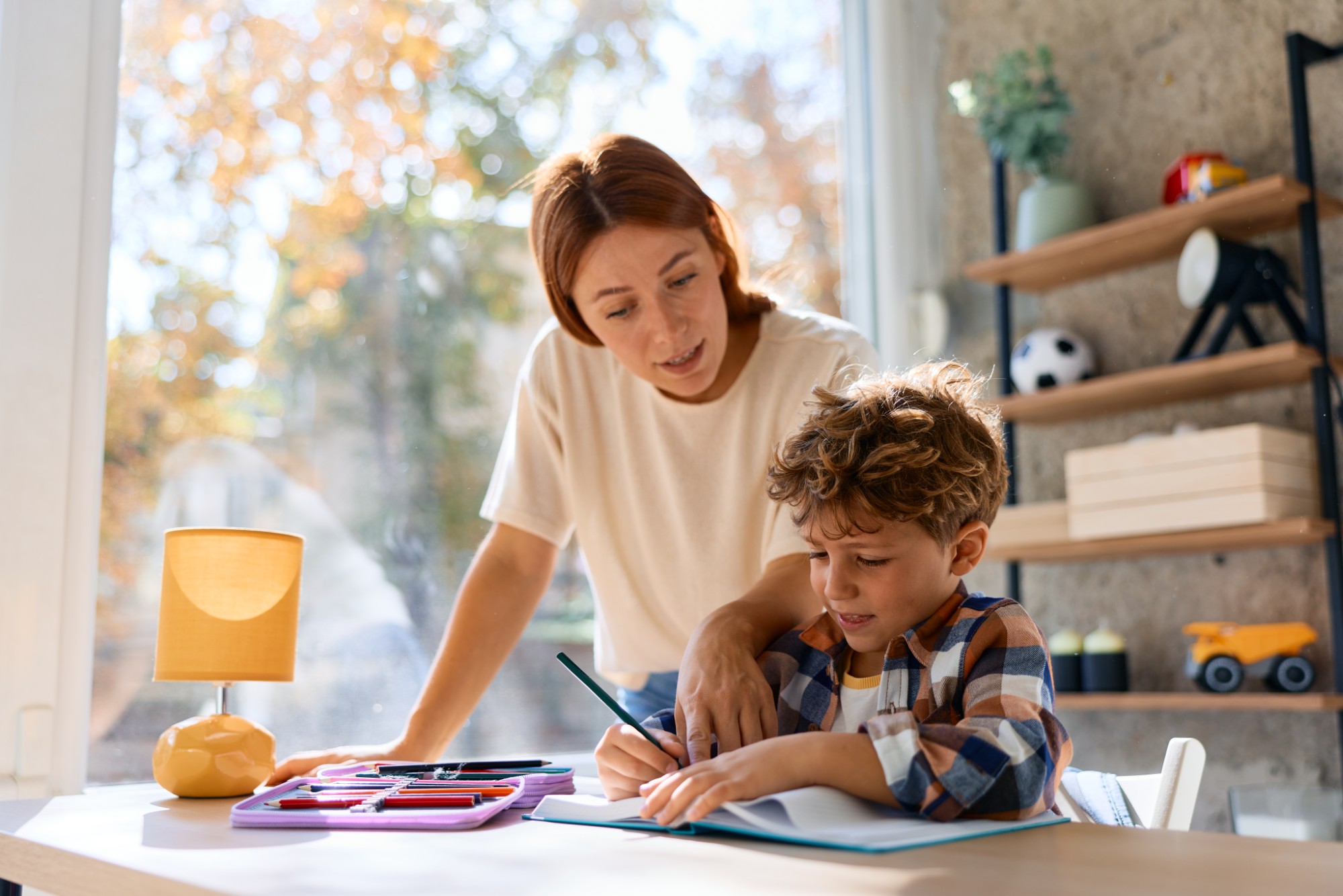 A woman helps a young boy with his homework at a desk by a window. Art supplies and a lamp are on the desk, and shelves with toys are seen in the background. Sunlight streams into the room.
