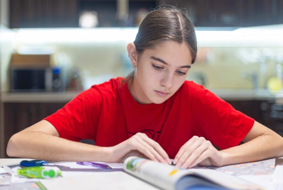 A young person with long hair, wearing a red shirt, sits at a table and focuses on reading an open book. School supplies, such as pencils and paper, are scattered on the table in a well-lit indoor setting.