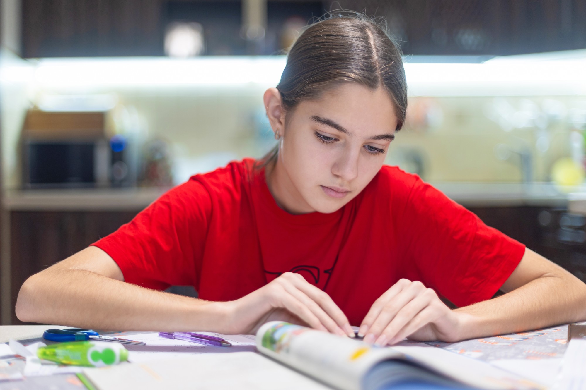 A young person with long hair, wearing a red shirt, sits at a table and focuses on reading an open book. School supplies, such as pencils and paper, are scattered on the table in a well-lit indoor setting.
