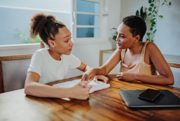 Two young women sit at a wooden table, engaged in conversation. One writes in a notebook while the other gestures supportively. A laptop and phone are on the table, and plants are visible in the background.