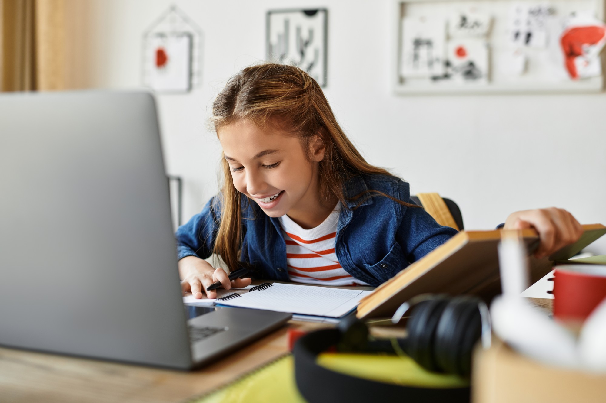A smiling young girl sits at a desk using a laptop, writing in a notebook. Art supplies, books, and headphones are on the desk. Childrens drawings hang on the wall in the background.