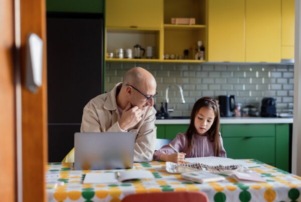 An older man and a young girl sit at a kitchen table with papers and a laptop, working together. The man looks at the girl, who is writing. The kitchen has yellow cabinets and a patterned tablecloth.