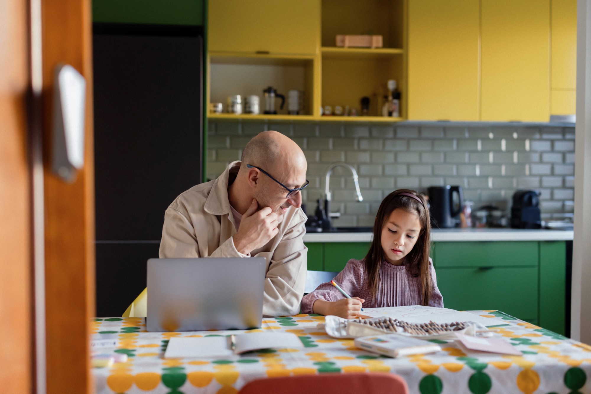 An older man and a young girl sit at a kitchen table with papers and a laptop, working together. The man looks at the girl, who is writing. The kitchen has yellow cabinets and a patterned tablecloth.