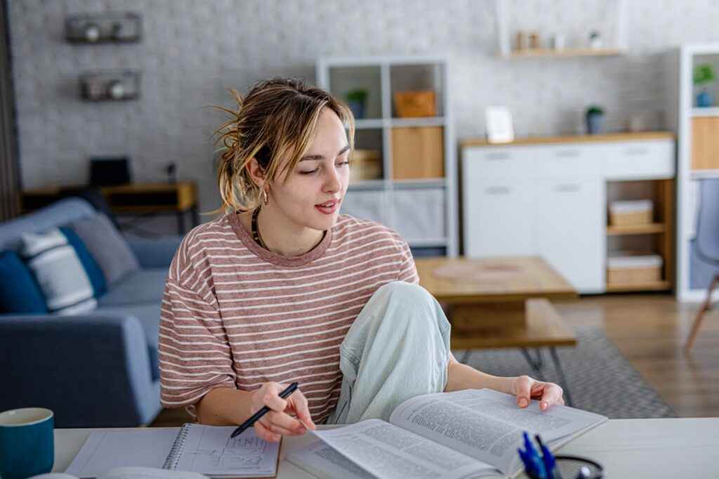 A young woman sits at a table in a living room, studying with open books and notes. She holds a pen and appears focused, with a cozy, modern background featuring shelves and a sofa.