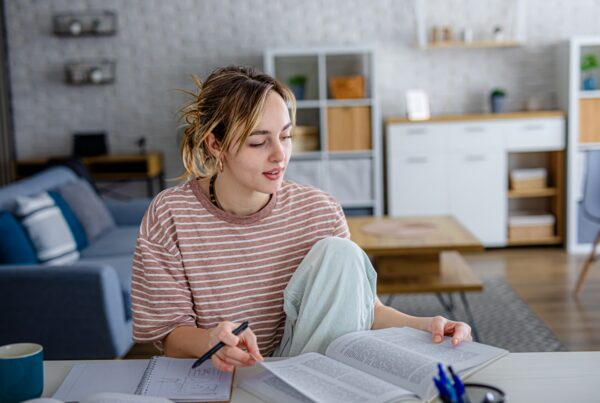 A young woman sits at a table in a living room, studying with open books and notes. She holds a pen and appears focused, with a cozy, modern background featuring shelves and a sofa.