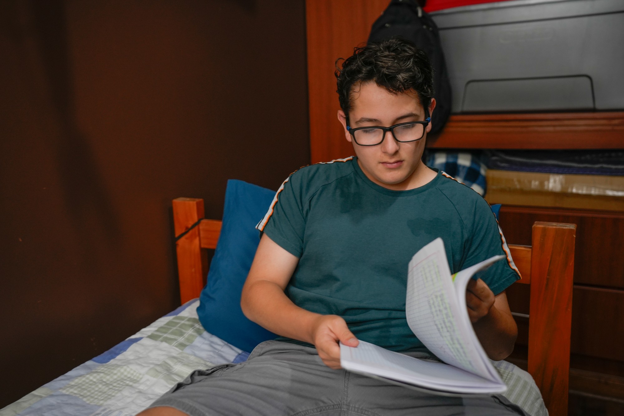 A teenage boy wearing glasses sits on a bed, reading or reviewing notes from a notebook. He is dressed casually in a green t-shirt and shorts, and the room has a wooden bed and shelves in the background.
