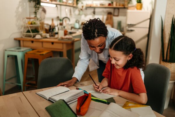 A woman helps a smiling girl with homework at a table in a cozy, well-lit kitchen. Books, notebooks, and pencils are spread out on the table around them.