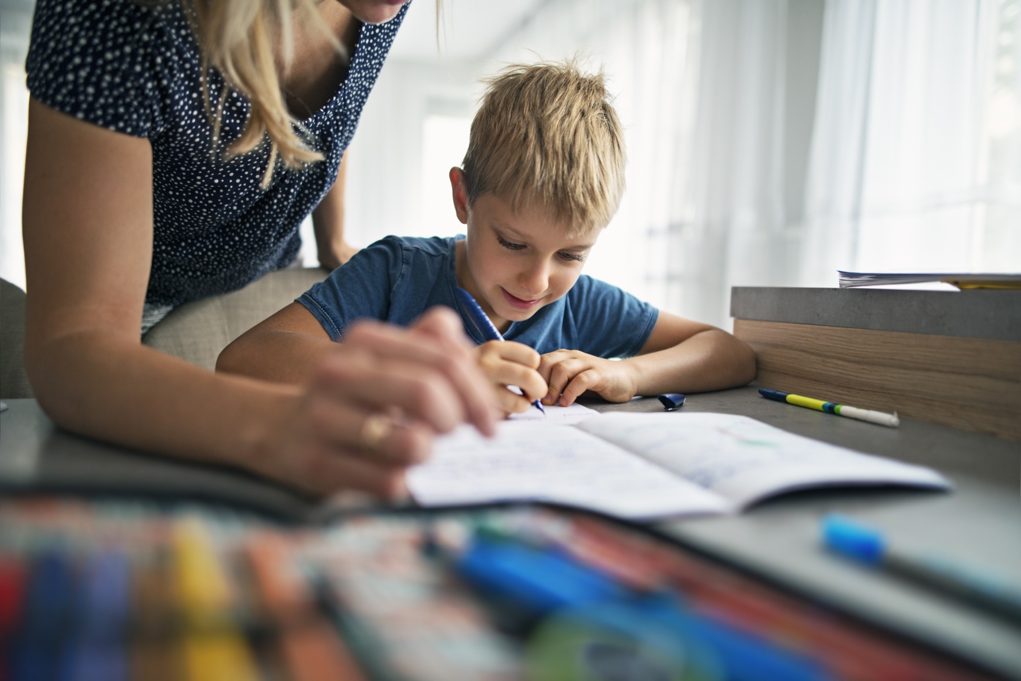 A young boy sits at a table, smiling and writing in a notebook, while an adult, partially visible, leans over to help. Colored pencils and markers are spread out on the table in the foreground.
