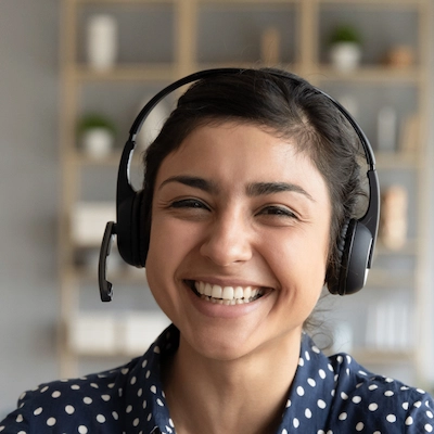 Smiling woman wearing a headset with microphone, dressed in a navy blue polka dot shirt, sitting indoors with blurred shelves and plants in the background.